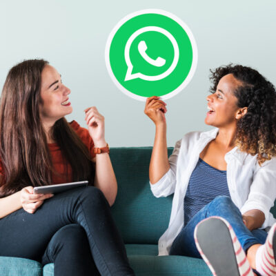 2 mulheres sentadas em um sofá conversando sobre como vender bem pelo WhatsApp Business. A moça da direita segura o simbolo do WhatsApp na mão.